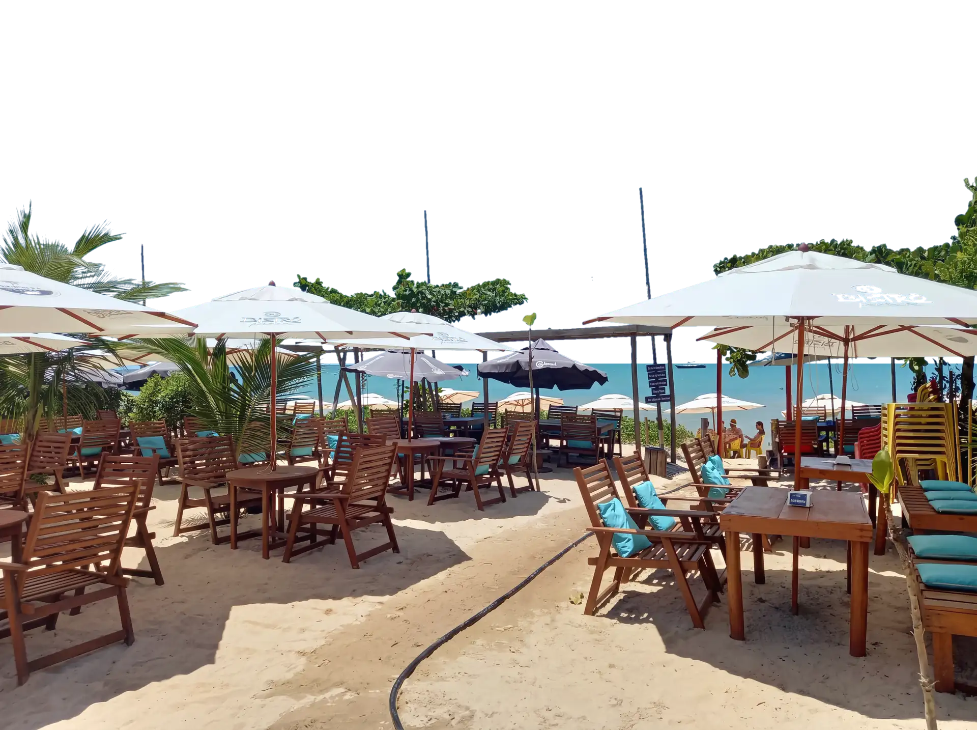 Mesas de madeira com almofadas azul-piscina na areia, guarda-sóis brancos e o mar